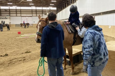 Volunteer Samantha Clark and another volunteer watch as client Rohan Kamath-Rayne receives help getting on the horse from his therapist.