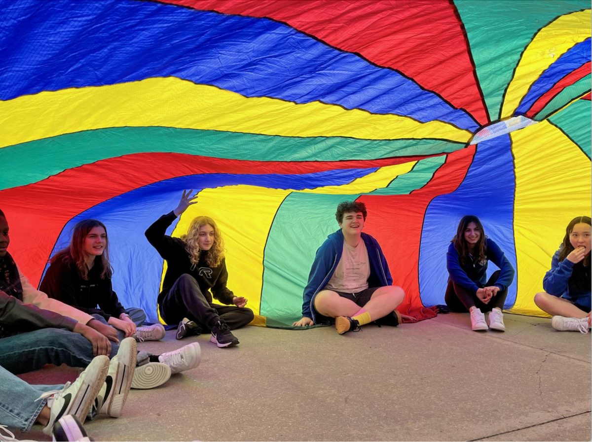 PEER-LEADING PURPOSE. (Left to right) Ninth graders Victoria Syverson and Paxton Cooper join junior peer leaders Theo Hinerfeld and Sofia Picciola under a parachute at the ninth grade energizer event on Apr. 9. The event, organized by the peer leader program and held on Kenwood Mall, offered different mini-games for ninth graders during Lab A. “I had a great time. It was a nice change to having to wake up and go straight to class and instead being able to go outside and get some sunlight,” ninth grader Isaac Sutherland said.