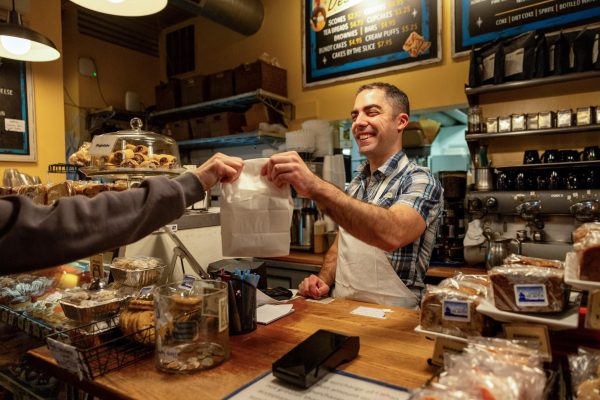 Behind the counter at A Taste Of Heaven in Andersonville, Zaccary Haney hands a bag of bakery items across the counter to the customer.
