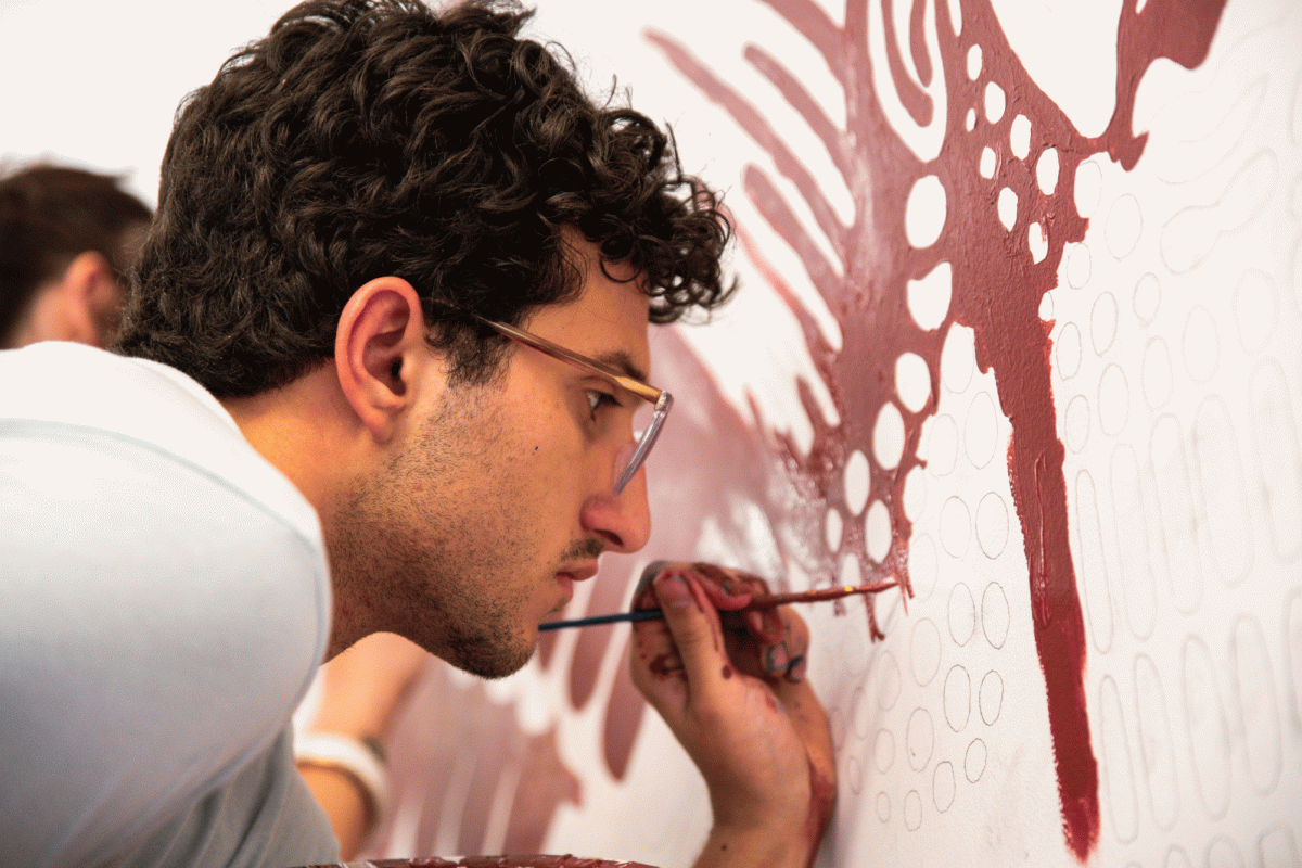 STEADY HAND. Holding his brush with careful concentration, senior Asher Dennis paints an intricate maroon design of a guardian, part of a larger mural outside of the U-High library. The mural was an hours-long project that lasted all day. 