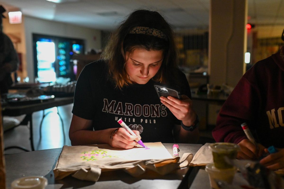 BRIGHT IDEAS. Peering down with intense focus, senior Emilia Anitescu uses her phone to illuminate a design she is creating on a tote bag, during Art in the Dark on Feb. 10. “I decorated a tote bag of things in my life that are artistic and things that I like, and added some pizazz to it to make it more fun,” Emilia said. “I also put a pin on it that I matched with my friends.”
